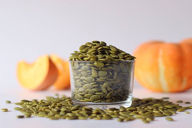Glass jar filled with green pumpkin seeds or pepitas on a white surface, surrounded by scattered seeds. Two orange pumpkins, one cut open, add autumn charm, ideal for healthy eating and food visuals